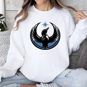 May include: White sweatshirt featuring a black bird design with outstretched wings, a red eye, and a blue star above. The bird is encircled by a blue and black design. The sweatshirt is a casual style.