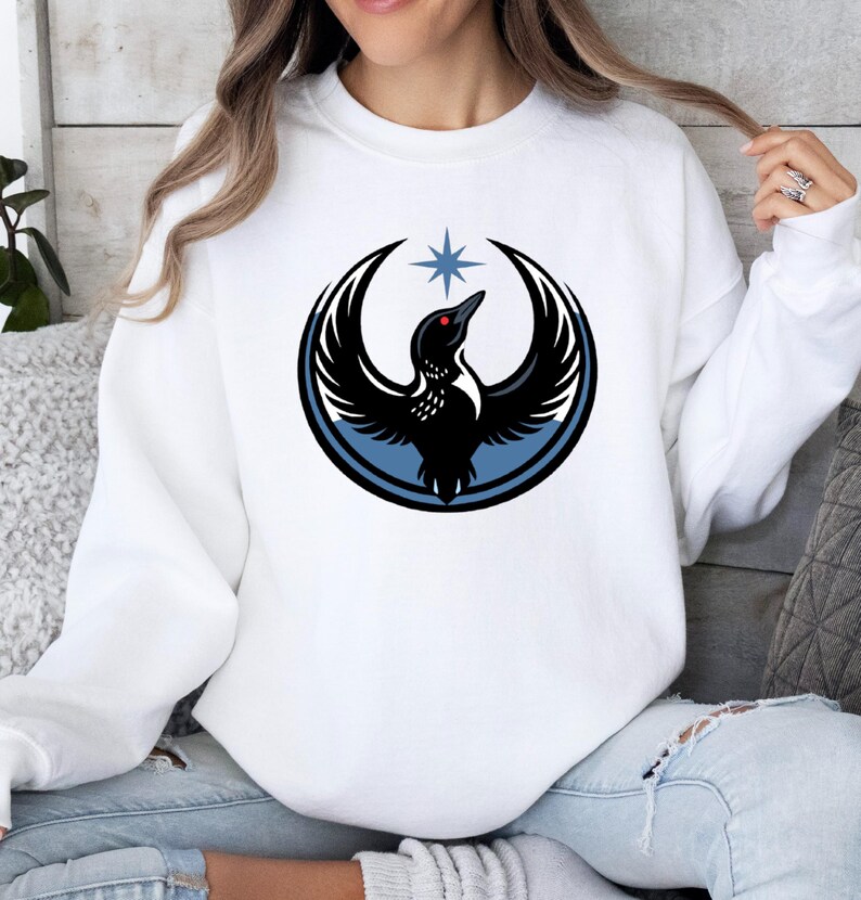 May include: White sweatshirt featuring a black bird design with outstretched wings, a red eye, and a blue star above. The bird is encircled by a blue and black design. The sweatshirt is a casual style.