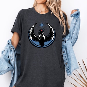 Puede incluir: Camiseta gris oscuro con un dise&ntilde;o circular de un p&aacute;jaro negro con las alas extendidas, un ojo rojo y una estrella azul encima. El p&aacute;jaro est&aacute; sobre un fondo circular azul y blanco. La camiseta tiene mangas cortas.