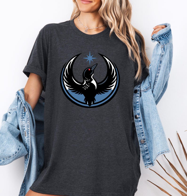 Puede incluir: Camiseta gris oscuro con un dise&ntilde;o circular de un p&aacute;jaro negro con las alas extendidas, un ojo rojo y una estrella azul encima. El p&aacute;jaro est&aacute; sobre un fondo circular azul y blanco. La camiseta tiene mangas cortas.