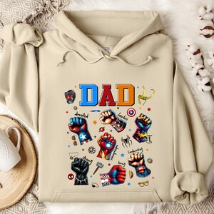 May include: A beige hoodie with the word "DAD" in colorful letters and a design of superhero fists with various words like "devoted", "fearless", "strong", "loving", "hardworking", and "funny" around them.