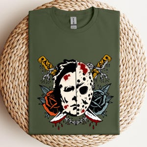 May include: Olive green t-shirt featuring a graphic design of a hockey mask split in half with two crossed knives. The design includes roses and blood splatter details, creating a horror-themed aesthetic.