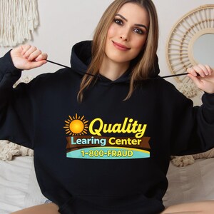 May include: Black hooded sweatshirt with the text "Quality Learing Center 1-800-FRAUD" in yellow, brown, and blue. The design includes a sun graphic. The sweatshirt is worn by a person.