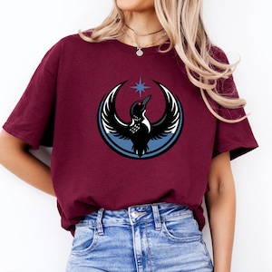 Puede incluir: Camiseta burdeos con un gr&aacute;fico de un p&aacute;jaro negro con detalles de alas blancas, un ojo rojo y una estrella azul. El p&aacute;jaro est&aacute; dentro de un dise&ntilde;o circular azul y blanco. La camiseta es de material suave.
