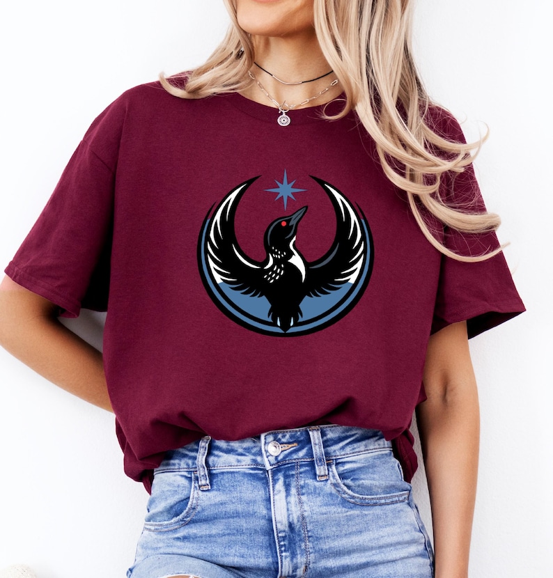 Puede incluir: Camiseta burdeos con un gr&aacute;fico de un p&aacute;jaro negro con detalles de alas blancas, un ojo rojo y una estrella azul. El p&aacute;jaro est&aacute; dentro de un dise&ntilde;o circular azul y blanco. La camiseta es de material suave.