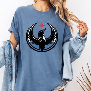 May include: A blue t-shirt featuring a black bird design with outstretched wings, a red star, and a blue circle. The bird has a white outline and a red eye. The shirt has rolled-up sleeves and a relaxed fit.