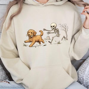 May include: A beige hooded sweatshirt featuring a graphic print of a golden-colored dog running with a bone in its mouth, followed by a human skeleton reaching out in a graveyard setting with a bare tree and a tombstone in the background.
