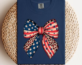 May include: Navy blue t-shirt featuring a patriotic bow design. The bow incorporates elements of the American flag with stars, stripes, and a leopard print pattern. The shirt is a Comfort Colors XL-TG-2X.