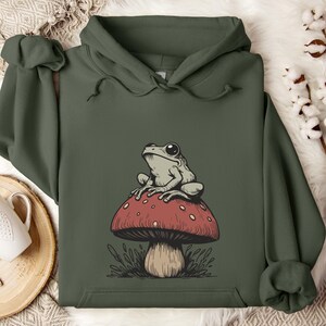 May include: A green hooded sweatshirt with a graphic of a frog sitting on a red and white mushroom. The mushroom has a brown stem and green leaves around the base.