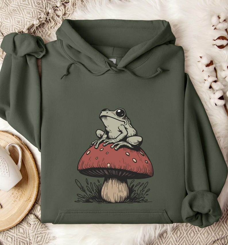 May include: A green hooded sweatshirt with a graphic of a frog sitting on a red and white mushroom. The mushroom has a brown stem and green leaves around the base.