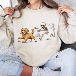 May include: A cream-colored crewneck sweatshirt featuring a graphic illustration of a golden-colored dog running with a bone in its mouth while being chased by a human skeleton in a graveyard setting with a bare tree and small tombstones.
