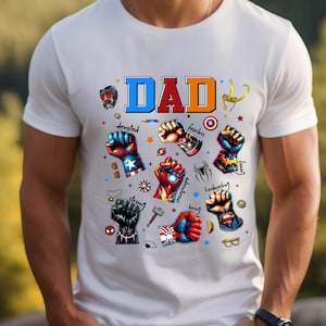 May include: White t-shirt with a colorful "DAD" graphic. The design features superhero fists with words like "devoted," "fearless," and "loving." The shirt has a crew neck and short sleeves.