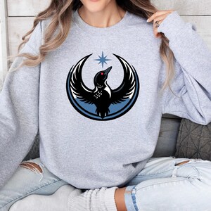 May include: A light gray sweatshirt featuring a circular graphic of a black bird with outstretched wings, a red eye, and a blue star above. The bird is set within a blue circle. The sweatshirt is worn by a person.