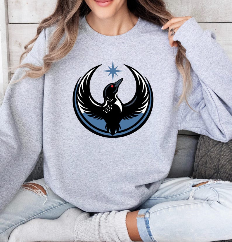 May include: A light gray sweatshirt featuring a circular graphic of a black bird with outstretched wings, a red eye, and a blue star above. The bird is set within a blue circle. The sweatshirt is worn by a person.