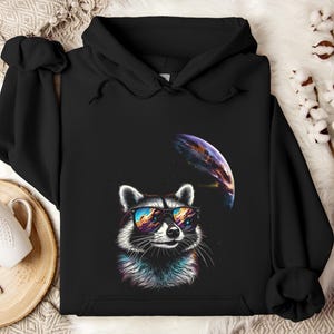 May include: Black hooded sweatshirt featuring a raccoon wearing sunglasses with a colorful sky reflection. A planet is in the background. The raccoon has a colorful fur design. The sweatshirt is made of a soft material.