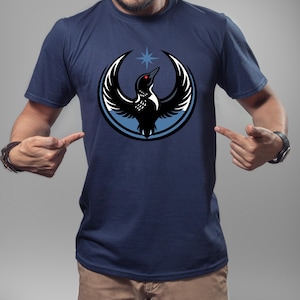 May include: Navy blue t-shirt featuring a stylized black bird with outstretched wings, a red eye, and a blue star above. The bird is encircled by a light blue ring and a white ring. The design is centered on the shirt.