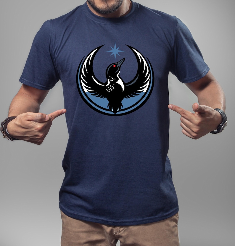 Puede incluir: Camiseta azul marino con un dise&ntilde;o de un p&aacute;jaro negro estilizado con alas extendidas, un ojo rojo y una estrella azul. El p&aacute;jaro est&aacute; rodeado por un c&iacute;rculo azul claro y blanco. El dise&ntilde;o est&aacute; centrado en la camiseta.