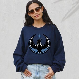 May include: Navy blue sweatshirt featuring a graphic of a bird with outstretched wings, a star, and a circular design. The bird is black and white with a red eye. The sweatshirt is a casual style.