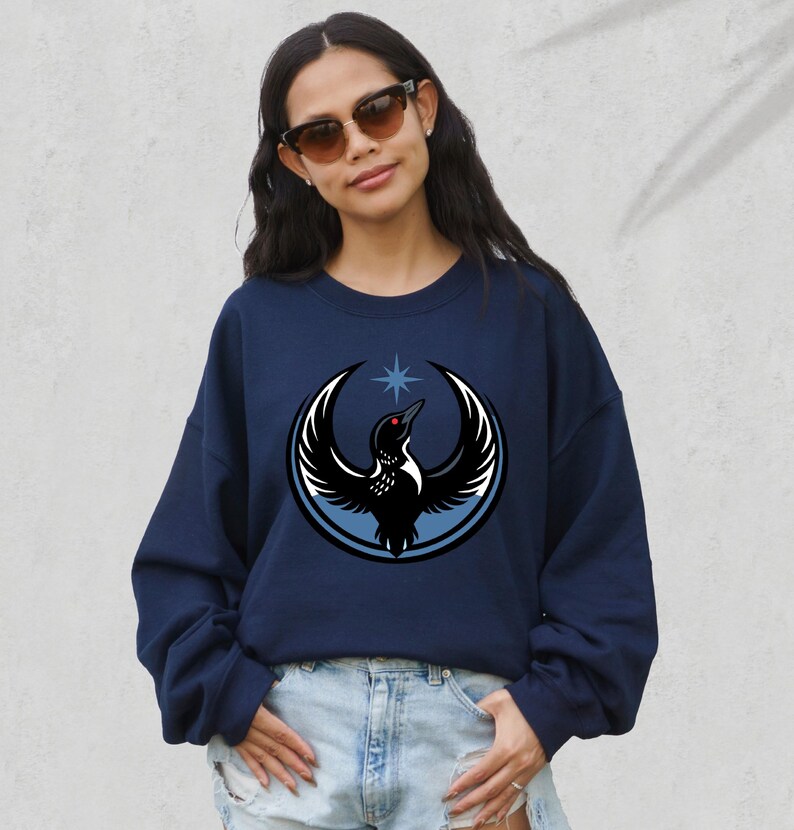 May include: Navy blue sweatshirt featuring a graphic of a bird with outstretched wings, a star, and a circular design. The bird is black and white with a red eye. The sweatshirt is a casual style.