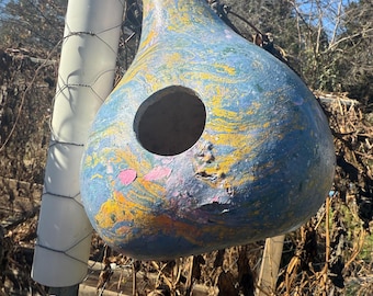 May include: A teardrop-shaped birdhouse with a large circular opening. The birdhouse is painted with a blend of blue, yellow, pink, and orange colors. It hangs from a wire and is set against a backdrop of trees and foliage.