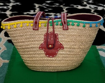 Puede incluir: Bolso tote de paja tejida con asas de cuero burdeos y un diseño de mano de Hamsa. El bolso presenta un ribete de pompón colorido en amarillo, verde y azul. El interior está forrado con tela burdeos.