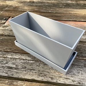 Könnte beinhalten: Ein rechteckiger grauer Pflanzer mit einem passenden Tablett. Der Pflanzer ist aus glattem Kunststoff.