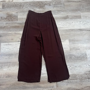 May include: High-waisted wide-leg trousers in a deep dark burgundy color. These pants feature a clean, minimalist waistband, subtle vertical pleats at the front, and a flowing, loose-fitting silhouette that extends down to the hemline.