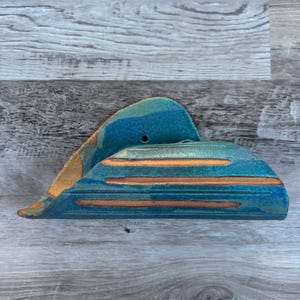 May include: A triangular ceramic napkin holder featuring a glossy teal and blue glaze with three horizontal, unglazed terracotta-colored stripes along the side. The piece has a small circular hole near the top edge and a smooth, curved form.