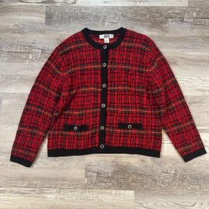 May include: A long-sleeve cardigan featuring a red and black plaid pattern with a black crew neckline, black button-down front, and two small front pockets. The garment has a black hem and cuffs, with a white label reading SEGUE at the neck.