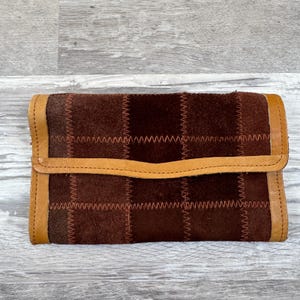 May include: A rectangular wallet constructed from dark brown suede patches joined by visible light brown zigzag stitching. The wallet features a smooth light brown leather trim along the edges and a horizontal flap closure across the center front.