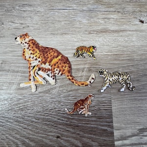 May include: Four embroidered patches of big cats. One large cheetah patch in shades of brown, tan, and white. Three smaller tiger patches, one with orange and black stripes, and two with brown and black stripes. All patches are on a wooden surface.