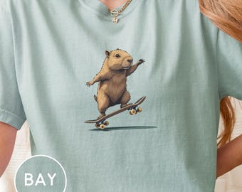 Puede incluir: Camiseta azul claro con un dibujo animado de una marmota en monopatín. La marmota es marrón con los brazos extendidos. El monopatín es marrón con ruedas amarillas. La palabra "BAY" está en un círculo blanco en la parte inferior izquierda.