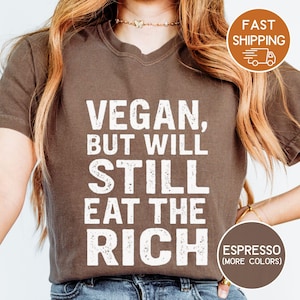 May include: Espresso-colored t-shirt with the text "VEGAN, BUT WILL STILL EAT THE RICH" in white block letters. The shirt has a crew neck and short sleeves. A round graphic indicates the shirt is available in more colors.