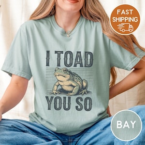 May include: A pale blue t-shirt featuring the phrase "I TOAD YOU SO" with a detailed illustration of a toad in the centre. The graphic is in grey and green tones. The shirt is a relaxed fit.