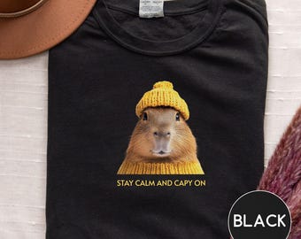 May include: Black t-shirt featuring a capybara wearing a yellow knit hat and sweater. The text "STAY CALM AND CAPY ON" is printed below the capybara. A black circle with the word "BLACK" is in the lower right corner.