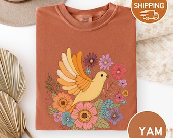 Könnte beinhalten: Rostfarbenes Rundhals-T-Shirt mit einem gelben Vogel mit orangefarbenen und gelben Flügeln, umgeben von bunten Blumen. Das Design umfasst orange, rosa, blaue und lila Blumen. Das Shirt hat den Text "Comfort Colors" auf dem Etikett.