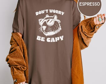 May include: Espresso-colored t-shirt with the text "DON'T WORRY BE CAPY" in white. The graphic features a capybara wearing sunglasses. The shirt is a relaxed fit, and the sleeves are rolled up. The model is wearing a brown jacket.
