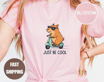 May include: A light pink t-shirt featuring a cartoon capybara wearing sunglasses and riding a teal scooter. The text "JUST BE COOL" is printed below the graphic. The shirt has a round neckline and short sleeves.