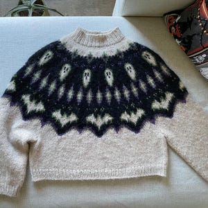 May include: A beige knit sweater featuring a dark black yoke with a repeating pattern of white ghosts and bats. The sweater has a high mock neck collar, long sleeves, and ribbed cuffs and hem, showcasing a detailed fair isle style design.