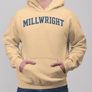 Op de afbeelding: Een lichtbeige hoodie met het woord "MILLWRIGHT" in marineblauw op de borst gedrukt. De hoodie heeft een voorzak en een capuchon. De persoon draagt een blauwe spijkerbroek.