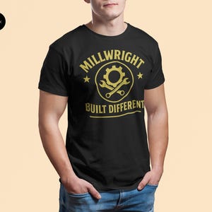 Op de afbeelding: Zwart T-shirt met een gouden opdruk met de tekst "MILLWRIGHT BUILT DIFFERENT". Het ontwerp bevat een tandwiel, een moersleutel en sterren. Het T-shirt heeft een ronde hals en wordt gedragen.