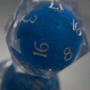 May include: Close-up of a clear, multi-faceted, twenty-sided die with a vibrant blue interior. The die features silver numerals, including the numbers 3, 8, 16, and 10. Ideal for tabletop gaming.