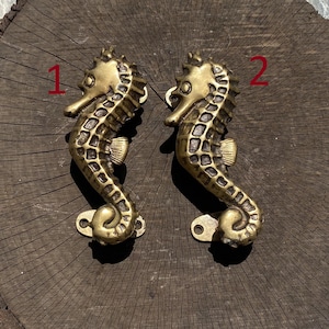 May include: Two gold-toned seahorse door handles, one labeled "1" and the other labeled "2", are displayed on a wooden surface.