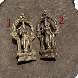 May include: Two antique brass statues of a female deity. Each statue is intricately detailed, featuring a figure within an arched frame. The figures are adorned with detailed clothing and jewelry. The statues are displayed on a weathered wooden surface.