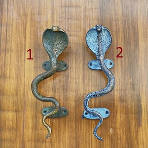 May include: Two ornate brass door handles shaped like cobras with their hoods raised. The handle on the left is a darker brass color, while the handle on the right is a lighter brass color with a blue patina.