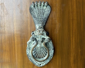 May include: A decorative door knocker made of metal with a green patina. The knocker is shaped like a serpent with a crown on its head. The serpent is holding a ring in its mouth.