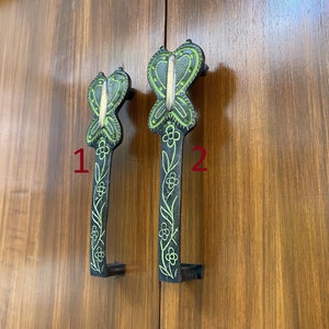 May include: Two black metal butterfly door handles with green floral details. The handles are mounted on a wooden surface. The handle on the left is labeled "1" and the handle on the right is labeled "2".