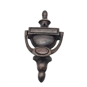 May include: A bronze-colored door knocker with a classic design. The knocker features a rectangular plate at the top, a rounded bowl shape in the middle, and a decorative base. The design includes rope-like detailing.