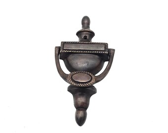 May include: A bronze-colored door knocker with a classic design. The knocker features a rectangular plate at the top, a rounded bowl shape in the middle, and a decorative base. The design includes rope-like detailing.