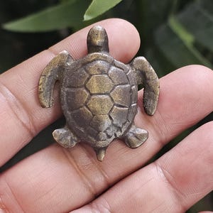Könnte beinhalten: Kleine Schildkrötenfigur in Bronze. Die Schildkröte hat ein detailliertes Schalenmuster und wird in einer Hand gehalten. Die Figur ist etwa 4 cm lang und hat eine detaillierte, strukturierte Oberfläche.